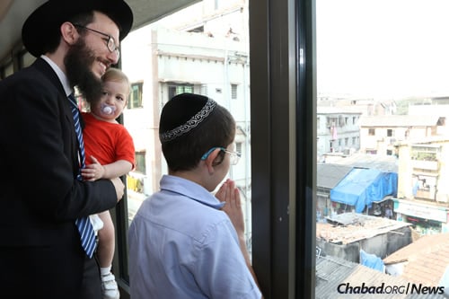 Moshe Holtzberg, right, fondly known as “Baby” Moshe, with Rabbi Israel Kozlovsky, co-director of Chabad-Lubavitch of Mumbai, and his baby, look out from Nariman House as well-wishers wave and blow kisses to Moshe. (Photo: Chabad of Mumbai/Chabad.org)