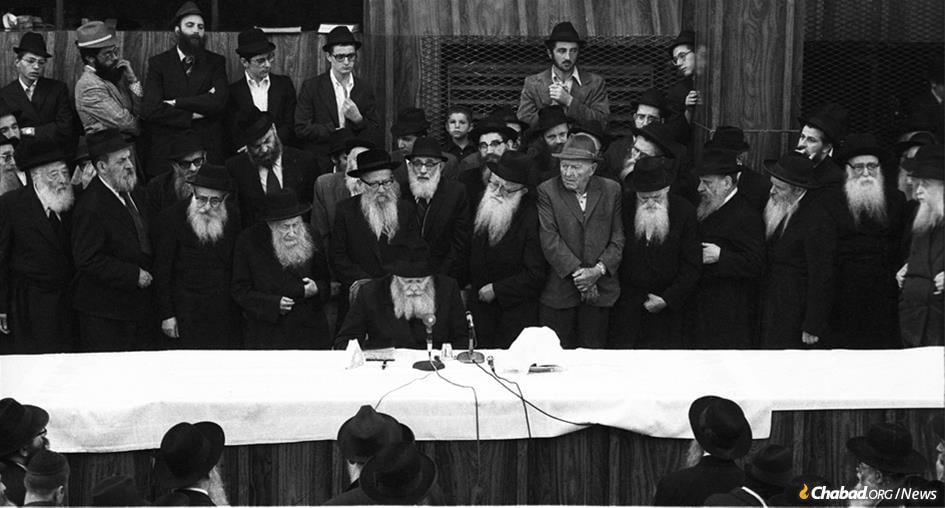The Rebbe recites a “maamar,” or Chassidic discourse, at a farbrengen gathering in the central Chabad synagogue at 770 Eastern Parkway in the Crown Heights neighborhood of Brooklyn, N.Y., on Sept. 4, 1975. While attendees mostly sat, the custom is to stand while the Rebbe teaches a discourse. The man wearing a hat in front of the Rebbe to his right is Rabbi Yoel Kahn, the Rebbe&#39;s chief chozer. (Photo: JEM/The Living Archive)
