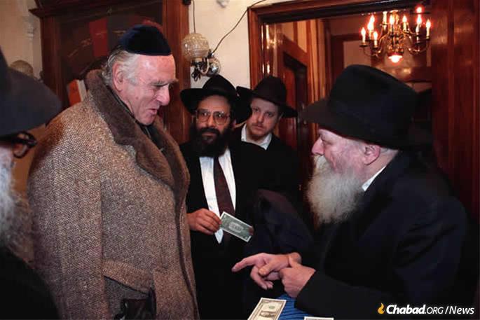 Federal Judge Jack B. Weinstein, then-chief and today senior judge on the U.S. District Court for the Eastern District of New York, receives a dollar and a blessing from the Rebbe—Rabbi Menachem M. Schneerson, of righteous memory—on Dec. 17, 1989, as Rabbi Sholom Lipskar, founder of the Aleph Institute, looks on. Weinstein was on his way to testify before the Federal Sentencing Commission and told the Rebbe he would be presenting the Rebbe&#39;s ideas before them. “You will support my views also, not just report them?“ the Rebbe asks, to which Weinstein responded he would. (Photo: Jewish Educational Media/The Living Archive)