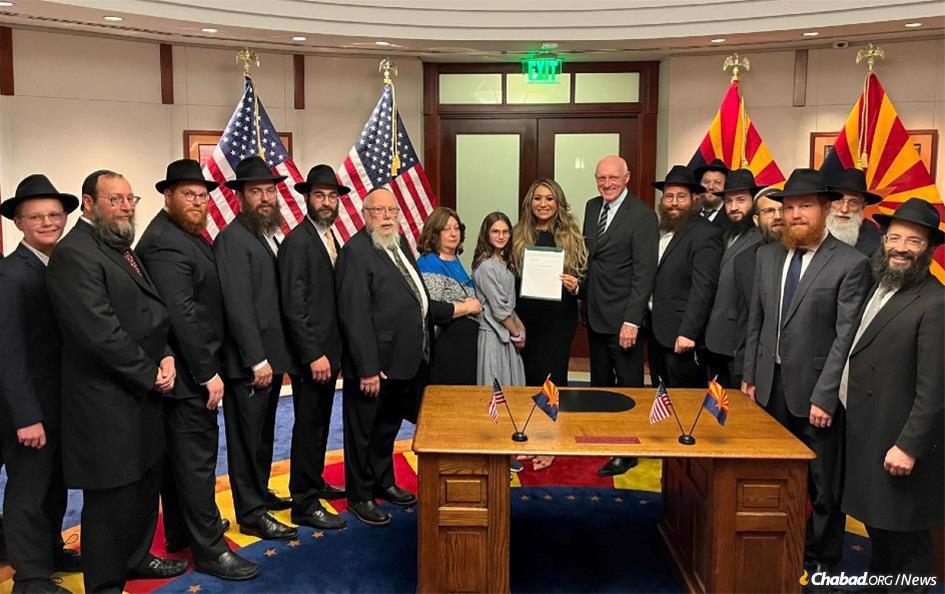 A Deputada Alma Hernandez (D-3º Distrito), centro, foi inspirada a fazer um Momento de Silêncio depois de conhecer o rabino Zalman Levertov,(sexto a partir da esquerda), diretor de Chabad-Lubavitch do Arizona, no acendimento da menorá em Chanucá. Eles são retratados aqui com a família Levertov, rabinos Chabad no Arizona e o governador do Arizona Doug Ducey na cerimônia de assinatura em Phoenix.