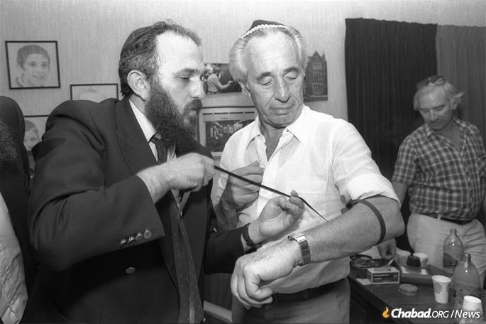 Chabad businessman Mayer Zeiler, who relocated to Israel at the Rebbe&#39;s behest in the 1970s, helps then-Prime Minister Shimon Peres don tefillin in 1985 at Zeiler&#39;s office in Nachalat Har Chabad. - Photo by Chananya Herman/GPO.