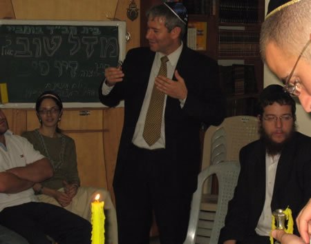 The Israeli Consul General in Mumbai addresses the celebration. Rivki and Gabi are seated to his right and left. The blackboard in the background reads: Chabad House of Mumbai. 20th of Elul. Mazal Tov to the Bar Mitzvah boy Ziv Sinai. All are invited! Mitzvah feast tonight at 19:00