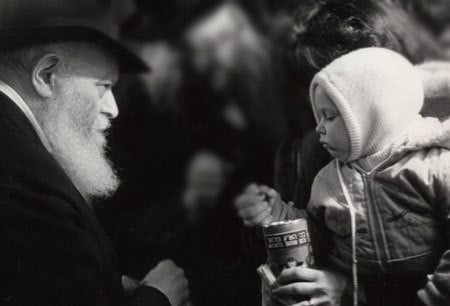The Rebbe waits as a child places a coin that the Rebbe just gave him into a charity box that his mother is holding. (Photo: Algemeiner Journal/Lubavitch Archives)
