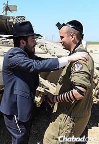 Cunin wraps tefillin with an Israeli soldier, surrounded by tanks and other military equipment. (Photo: Meir Alfasi)